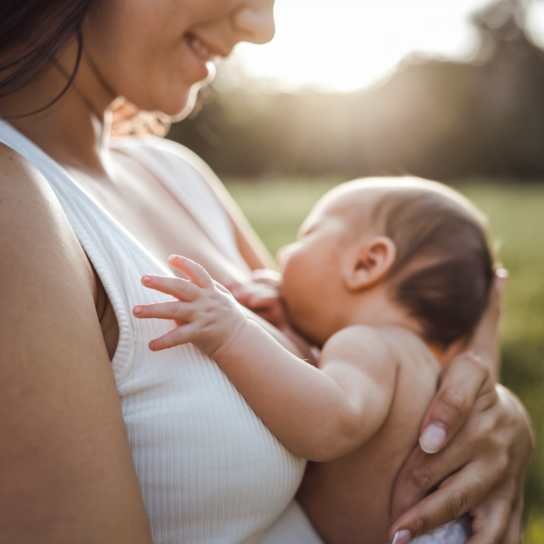 A parent smiles while breastfeeding their baby outdoors in a sunlit area.